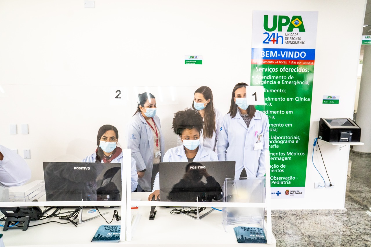 FOTO DE RECEPÇÃO -  Atrás de um balcão branco temos, quatro profissionais uniformizadas, duas sentadas e duas em pé trabalhando, na parede branca se destaca a placa colorida que traz o nome da UPA